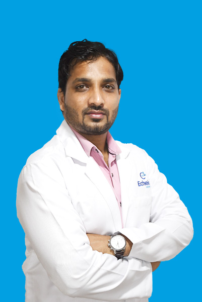 Male doctor in a white coat with pink shirt, arms crossed against a blue studio backdrop.