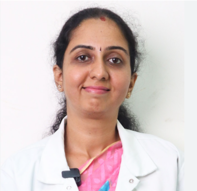 Portrait of a smiling woman in a white lab coat and pink blouse, wearing a bindi, against a light background.