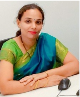 Professional woman in a green and blue sari seated at a desk with a computer mouse, looking at the camera.