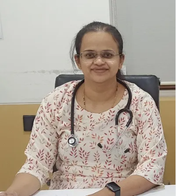 Smiling female doctor with a stethoscope around her neck, seated at a desk in a clinical office wearing glasses and a patterned blouse.