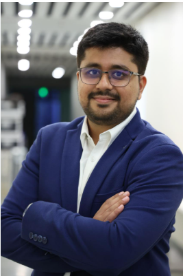 Professional portrait of a man in a blue blazer and white shirt, with glasses, arms crossed in a bright hallway.