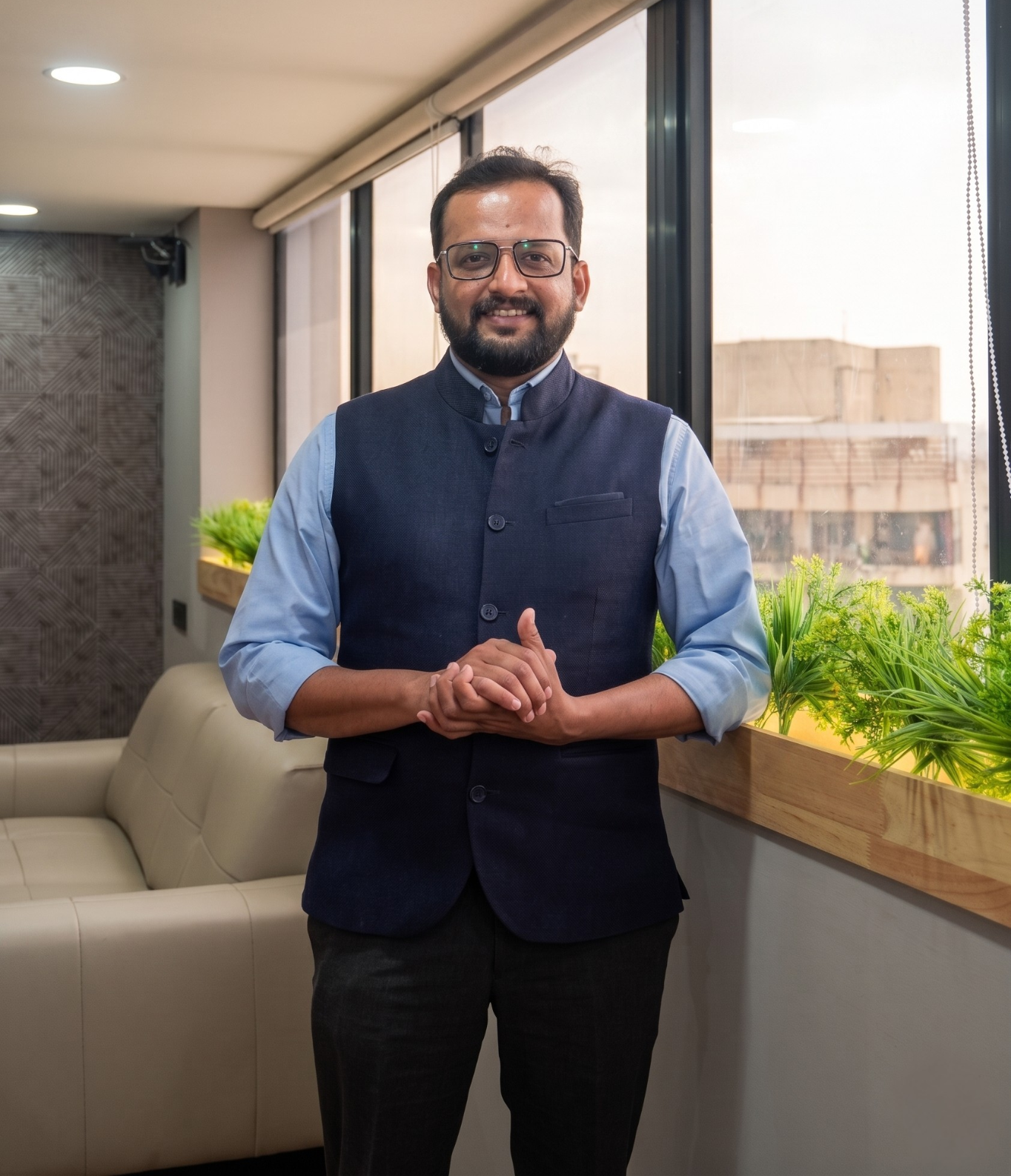 Man with glasses and a trimmed beard, wearing a blue shirt and navy vest, standing with hands clasped in a bright office lounge beside windows and plants.