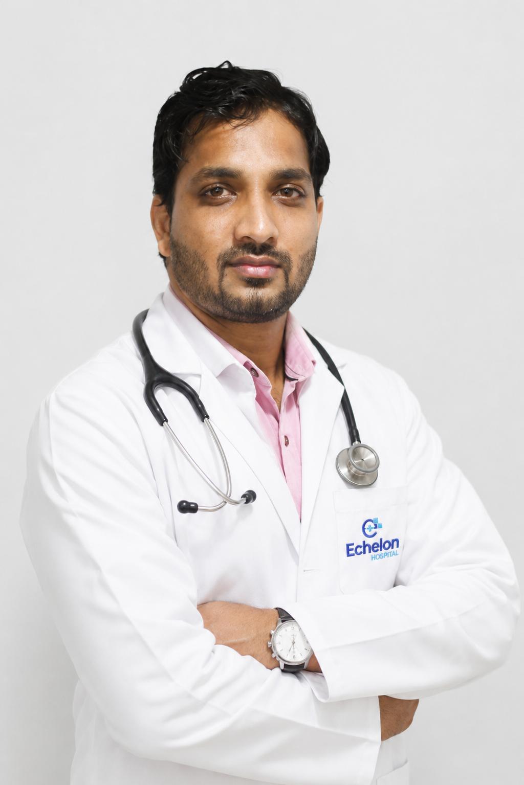 Portrait of a male doctor in a white coat with a stethoscope, arms crossed, wearing a pink shirt, Echelon Hospital logo on the coat