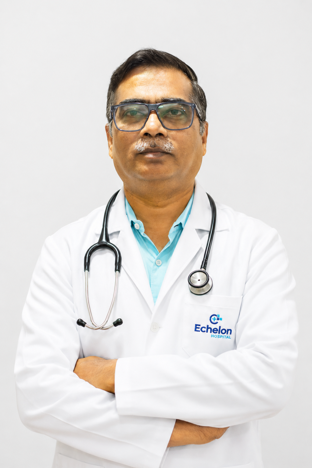Male doctor in a white coat with stethoscope, arms crossed, wearing glasses, against a light background with Echelon Hospital logo on the pocket.