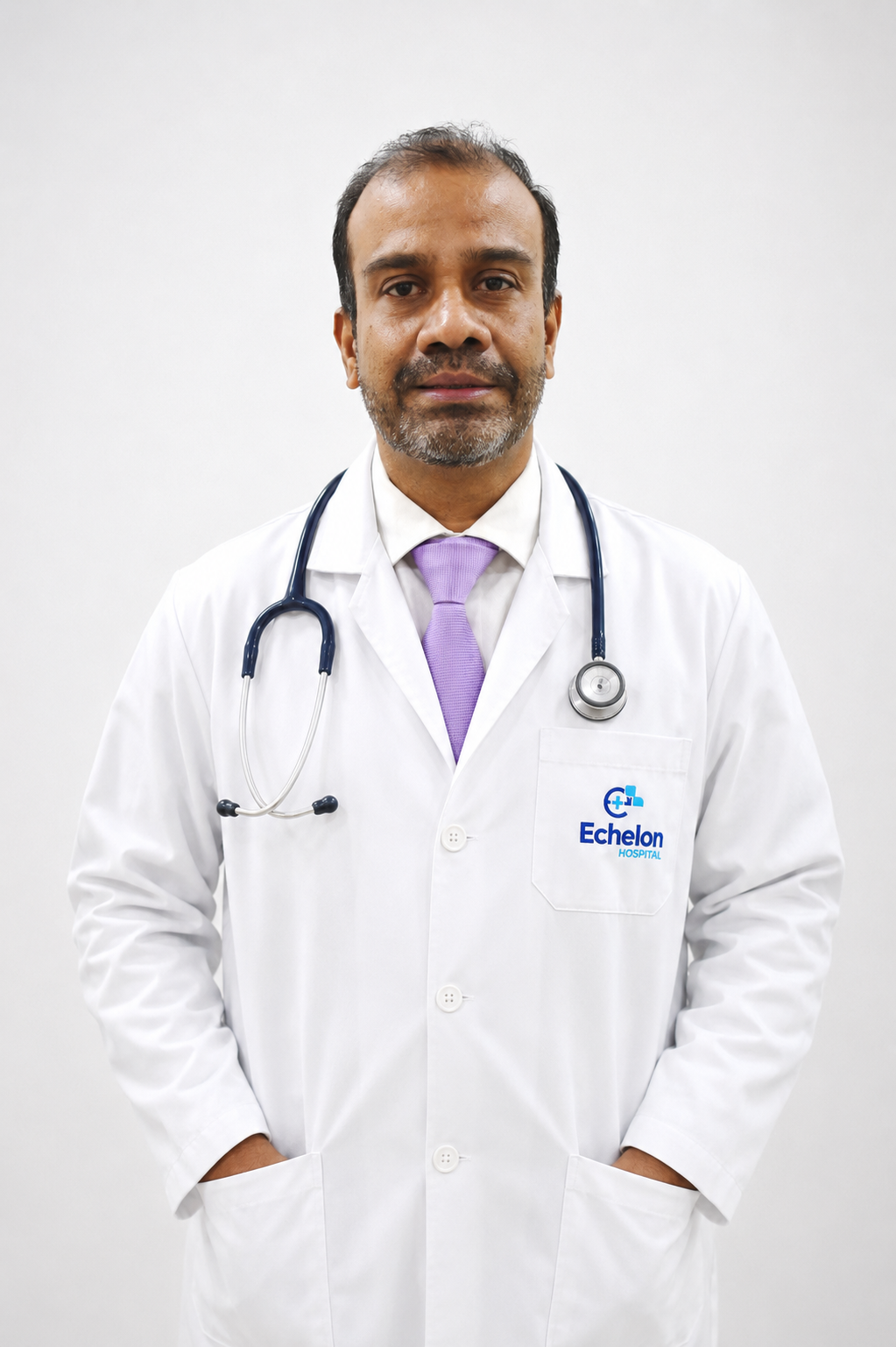 Male doctor in a white coat with a stethoscope around his neck, lavender tie, hands in pockets, Echelon Hospital logo on the chest pocket.