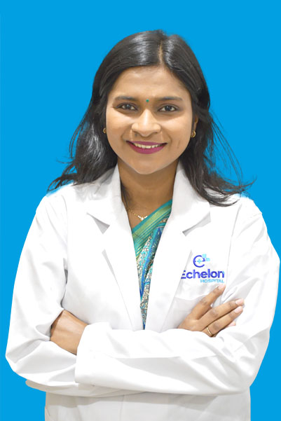 Female doctor with arms crossed, wearing a white coat over a teal outfit, standing against a blue background with a hospital logo on the coat.
