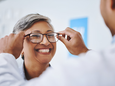 Optometrist or clinician adjusting a woman's glasses as she smiles in a clinical setting.