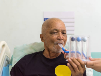 Older man in a hospital bed drinks through a blue straw while a clinician holds a rack of four clear test tubes beside him.