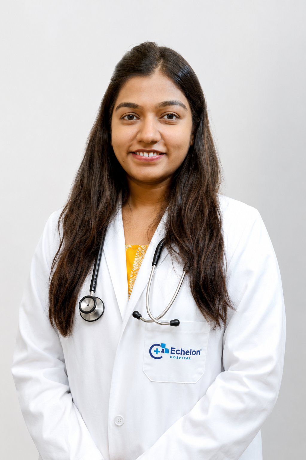 Smiling female doctor with long dark hair wearing a white coat and stethoscope, standing against a light background at Echelon Hospital.