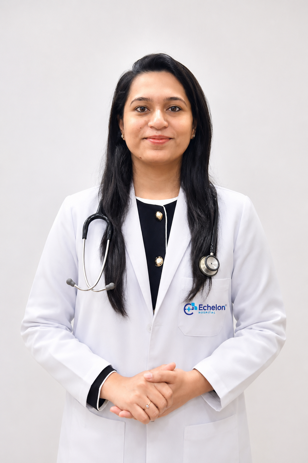 Female doctor in a white coat with stethoscope around her neck, standing with hands clasped; Echelon Hospital logo on the pocket.