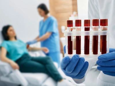 Gloved hands hold a rack of blood sample tubes in a clinical setting, with a patient and nurse in the background.