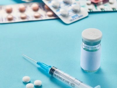 Medical supplies on a blue surface: a syringe, a small vial, and scattered pills with blister packs in the background.