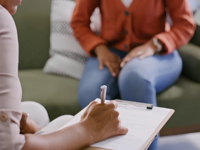 Person writes on a clipboard held on their lap during a seated session with another person in the background on a couch